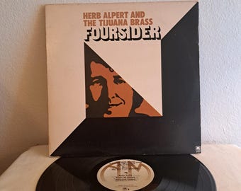 May include: A black vinyl record with a white label featuring the text "Herb Alpert and The Tijuana Brass" and "Four Sider". The record is on a white surface with a black and white album cover behind it.