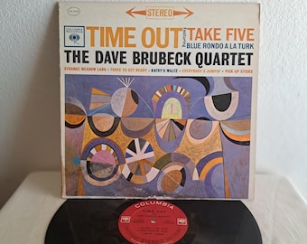 May include: A vintage vinyl record album cover featuring a colorful abstract design with circles and lines in shades of blue, yellow, red, and white. The album is titled "Time Out" by "The Dave Brubeck Quartet" and features the song "Take Five" with the subtitle "Blue Rondo A La Turk".