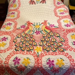 May include: A white crocheted bedspread with a pink and green floral design featuring two peacocks. The peacocks have yellow, red, and green feathers.
