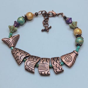 May include: Copper-toned fish-themed bracelet with a lobster clasp closure. The bracelet features fish-shaped charms, green, purple, and gold beads. The bracelet is approximately 7 inches long.