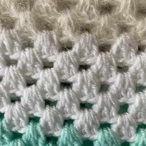 Peut inclure: Gros plan sur une couverture au crochet avec trois bandes de couleurs différentes. La bande supérieure est beige clair, la bande du milieu est blanche et la bande inférieure est vert menthe clair.