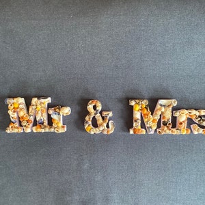 May include: White letters spelling "Mr. & Mrs." decorated with small seashells in shades of brown, beige, and purple.