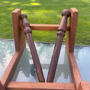 May include: Two wooden dowels with rounded ends are displayed in a wooden stand. The stand is made of a reddish-brown wood and the dowels are a darker brown.