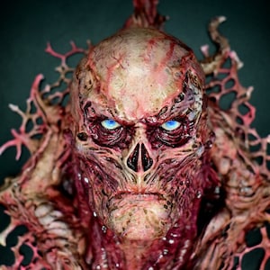 Könnte beinhalten: Eine detaillierte, groteske Maske mit einem verfallenden, fleischartigen Aussehen. Die Maske weist freiliegendes Gewebe, eine markante Nase und stechende blaue Augen auf. Das Design umfasst komplizierte, venenartige Strukturen und einen dunklen Hintergrund, wodurch ein Horror-Thema entsteht.