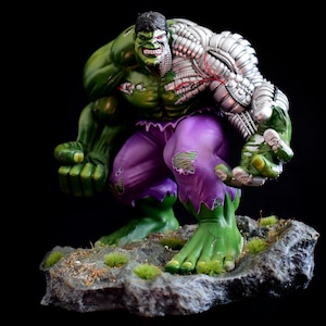 Peut inclure: Une figurine d'action verte de Hulk avec un bras robotique argenté, portant un pantalon violet déchiré, debout sur une base rocheuse avec de la mousse verte.