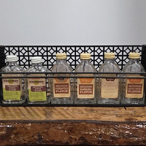 Puede incluir: Un estante de metal negro vintage sostiene seis botellas de vidrio con etiquetas variadas, incluyendo "Imitation Maple Flavor" y "Extract of Orange". Las botellas tienen tapas blancas o amarillas y están sobre una superficie de madera.