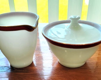 May include: A white creamer and sugar bowl set with a red and gold band around the top. The creamer has a pouring spout and the sugar bowl has a lid.
