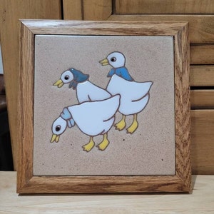 May include: A ceramic tile with a wooden frame features a whimsical illustration of three white ducks with blue accents. The ducks are depicted in a playful pose, with one duck looking back at the viewer.