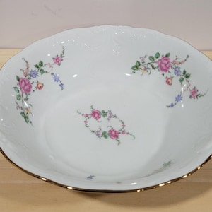 May include: A white ceramic bowl with a gold rim and a floral design. The bowl has a scalloped edge and features pink, blue, and green flowers.