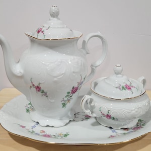 May include: A white porcelain tea set with floral designs. The set includes a teapot, creamer, and a tray.
