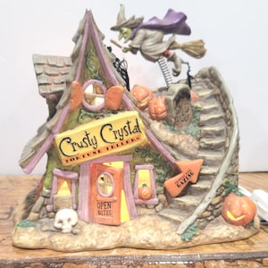 May include: A whimsical Halloween-themed ceramic figurine of a fortune teller's shop. The shop is adorned with pumpkins, a witch on a broom, and the sign "Crusty Crystal Fortune Tellers". The shop has an open door with the text "Open Nites".