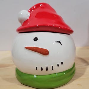 Op de afbeelding: Witte keramische sneeuwpopfiguur met een rode kerstmanmuts, een groene sjaal en een knipperend gezicht. De sneeuwpop heeft een wortelneus en zwarte ogen.