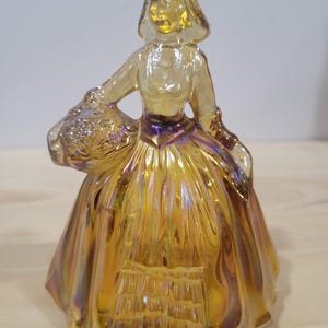 Puede incluir: Una figurita de vidrio de una mujer con un vestido amarillo iridiscente que sostiene una cesta de flores. La figurita está hecha de vidrio de carnaval y tiene un aspecto vintage.