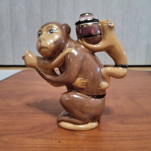 May include: A ceramic figurine of two monkeys, one carrying a small, ornate pot on its back. The monkeys are brown and have painted details, including eyes and a black band around the waist of the larger monkey.