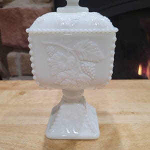 May include: A white, lidded candy dish with a square base and a decorative grape design. The dish is made of opaque glass and has a beaded edge. The lid has a small, decorative handle.