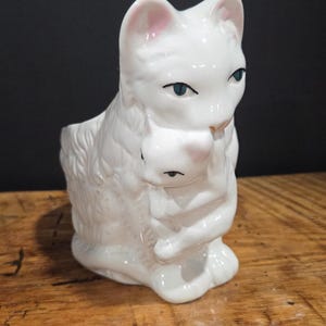 May include: White ceramic cat figurine with a mother cat embracing a kitten. The figurine has blue eyes and pink accents on the ears and nose. The cat is sitting on a wooden surface.