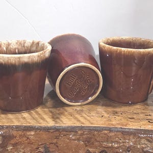 Op de afbeelding: Drie bruine keramische mokken met een glanzende afwerking en een crèmekleurig druipglazuur rond de bovenranden. Elke mok heeft een cirkelvormig handvat. De onderkant van de middelste mok is zichtbaar met de tekst "hull oven proof U.S.A."