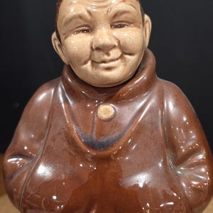 May include: A ceramic figurine of a smiling monk in a brown robe. The figurine has a detailed face with a bald head and a button on the robe. The base of the robe has a rope-like design.