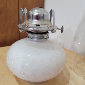 May include: A white glass oil lamp with a silver metal top and a textured surface. The lamp has a round shape and a small knob on the side.