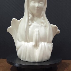 Puede incluir: Un busto de cerámica en color blanco roto mate que representa una figura con un velo sobre la cabeza y las manos juntas en posición de oración. La figura se muestra desde el pecho hacia arriba, con contornos suaves y redondeados.