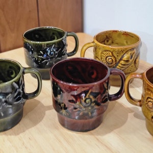 Puede incluir: Conjunto de cuatro tazas de cerámica con un diseño vintage. Cada taza presenta un patrón diferente de conchas marinas y otros motivos inspirados en el océano. Las tazas son en tonos de verde, marrón y amarillo.