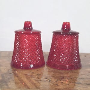 May include: A pair of matching ruby red glass shakers featuring a textured diamond pattern across the exterior. Each shaker has a tapered cylindrical shape with a flat top and a small central opening, showcasing a translucent finish throughout.
