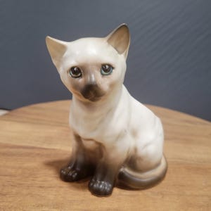Op de afbeelding: Een keramiek beeldje van een wit Siamese kitten met blauwe ogen, zittend met zijn pootjes ingetrokken. Het beeldje is geschilderd met realistische details, waaronder een lichtbruine neus en snorharen.