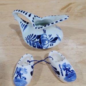 May include: A set of three miniature Dutch Delftware items: a blue and white pitcher with a floral design, and two blue and white clogs with a windmill design. The clogs are connected by a blue string.