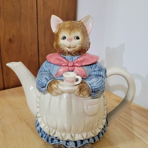 Puede incluir: Una tetera de cerámica con forma de gato que lleva un suéter azul, un lazo rosa y un delantal blanco. El gato sostiene una taza de té pequeña.
