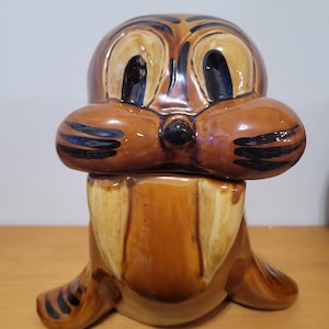 May include: A brown ceramic figurine of a cartoon walrus with black stripes and blue eyes. The walrus has a large, open mouth with two long, white tusks.