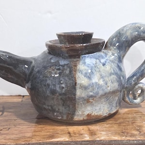 May include: A handcrafted ceramic teapot with a unique design. The teapot features a two-tone colour scheme, with a dark grey and blue-grey glaze. The handle has a curled end. The lid is a dark brown colour. The teapot is sitting on a wooden surface.