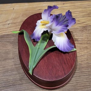 May include: A decorative iris flower sculpture with purple and white petals, a yellow center, and green leaves. The flower is mounted on a dark red, oval-shaped wooden base.