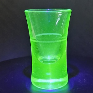 May include: A vibrant neon green shot glass made of uranium glass, which glows under ultraviolet light. The glass features a smooth, slightly flared cylindrical shape with a thick, solid base and a subtle horizontal etched line around the middle.