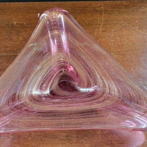 May include: A triangular, pink glass dish with a swirling, textured design. The dish has a rounded shape and is made of transparent glass. The color is a soft pink with hints of gold, creating a decorative piece.
