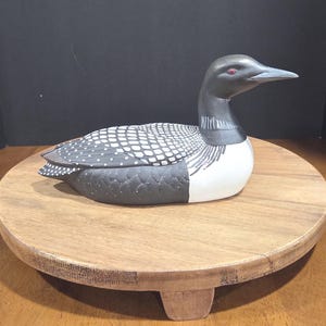May include: A hand-carved wooden loon figurine with a black head, white and black speckled back, and white underbelly. The bird has a red eye and a gray beak. It rests on a round, wooden pedestal.