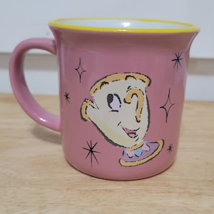May include: A pink ceramic mug with a yellow rim features a cartoon illustration of Lumiere, the animated candlestick from Disney's Beauty and the Beast. The illustration shows Lumiere's face and body, with a yellow and blue design on his collar.