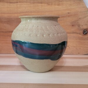 May include: A beige ceramic vase with a rounded body and a narrow neck. The vase features a horizontal band of dark teal and maroon paint. Decorative etched lines encircle the top. The vase is displayed on a wooden surface.