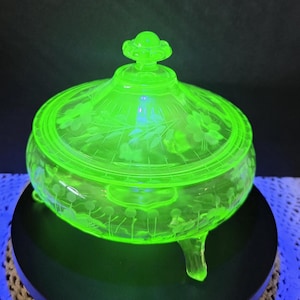 May include: A glowing neon green glass candy dish with a matching lid, featuring an etched floral pattern and delicate leaf designs. The circular vessel stands on three small glass feet and is topped with a decorative knob handle.