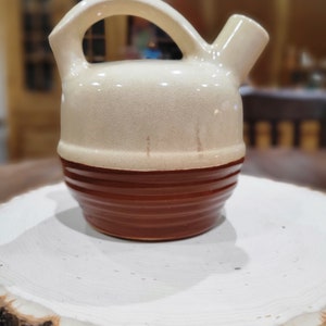 May include: A ceramic pitcher featuring a cream-coloured upper section with a subtle crackle glaze and a dark brown ribbed lower base. The vessel includes a curved handle extending from the top and a short, angled spout for pouring liquids.