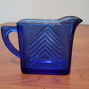 May include: A cobalt blue glass creamer with a geometric design. The creamer has a square body and a spout.