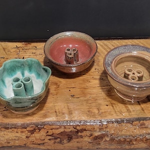 Könnte beinhalten: Drei Keramik-Blumenfrösche in Grün-, Rot- und Beigetönen. Jeder Frosch hat eine kreisförmige Schale mit drei Löchern zum Anordnen der Stiele. Die Frösche werden auf einer rustikalen Holzoberfläche ausgestellt.
