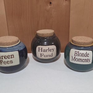 May include: Three round ceramic jars with cork lids. The left jar is dark blue with the text Green Fees. The center jar is black with the text Harley Fund. The right jar is teal with the text Blonde Moments. Each jar features a white label.