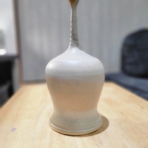 Puede incluir: Un jarrón de cerámica blanco con un cuello alto y delgado y un cuerpo redondeado y bulboso. El jarrón tiene un esmalte marrón claro y una superficie ligeramente texturizada.