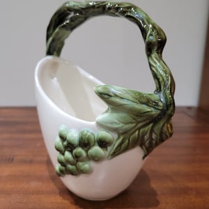 May include: A white ceramic planter shaped like a basket with a green vine handle and green grapes. The planter is decorated with green leaves.