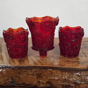 Puede incluir: Tres portavelas votivos de vidrio rojo rubí con un patrón floral en relieve y bordes festoneados. El portavelas central está elevado sobre una pequeña base de pedestal, mientras que los dos laterales tienen bases planas, todos con acabado translúcido.