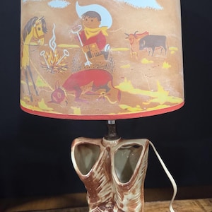 Może przedstawiać: Lampa w stylu vintage z motywem kowbojskim. Beżowy abażur przedstawia scenę kowbojską z ogniskiem, koniem i bydłem. Podstawa ma kształt brązowych ceramicznych butów kowbojskich. Abażur ma czerwone obramowanie.