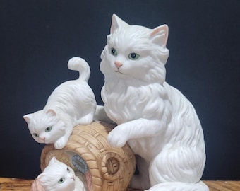 Puede incluir: Figura de cerámica blanca de una familia de gatos. Se representa una gata madre y dos gatitos, uno sobre una cesta de mimbre y otro dentro. Los gatos tienen ojos verdes y están sobre una base de madera.