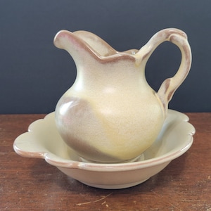 Pode incluir: Um conjunto de jarro e bacia de cerâmica. O jarro tem uma alça e um bico curvos, com um corpo arredondado. A bacia tem uma borda ondulada. O conjunto é principalmente bege com detalhes em marrom e rosa, sugerindo uma estética vintage.