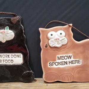 Puede incluir: Dos placas de cerámica con forma de gato. Una es negra con el texto "MOUSEWORK DONE FOR FOOD". La otra es de color melocotón con el texto "MEOW SPOKEN HERE". Ambas tienen una cuerda marrón para colgar y detalles de cara de gato.