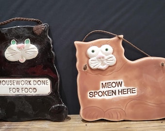 May include: Two ceramic cat-shaped plaques. One is black with the text "MOUSEWORK DONE FOR FOOD". The other is peach-colored with the text "MEOW SPOKEN HERE". Both have a brown rope for hanging and cat face details.